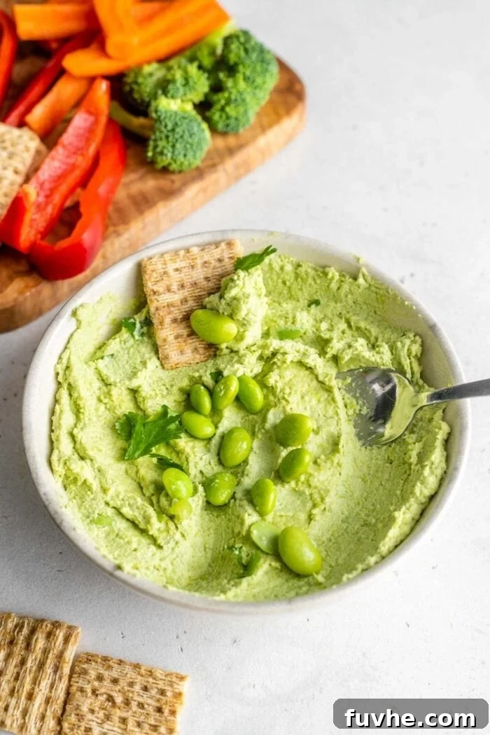 A spoon and cracker scooping edamame hummus out of a small bowl sitting on a white surface, showcasing its smooth texture.