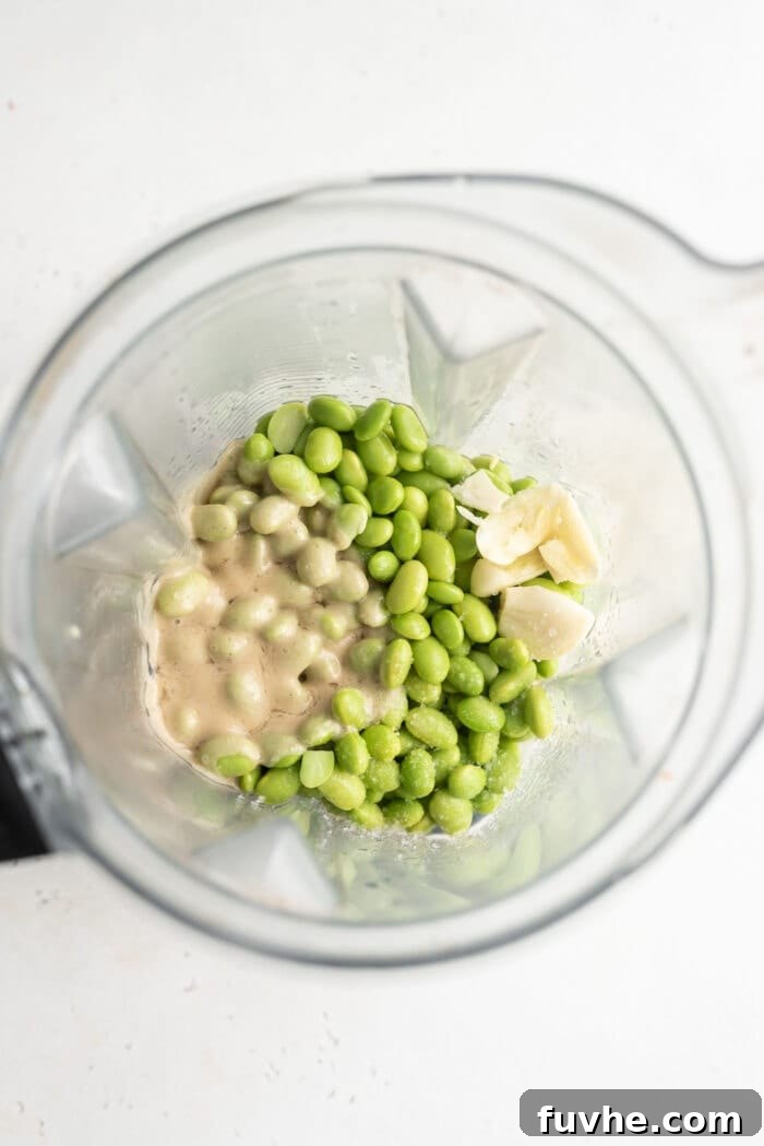 Overhead view into a blender container with edamame, tahini, and garlic, prior to blending.