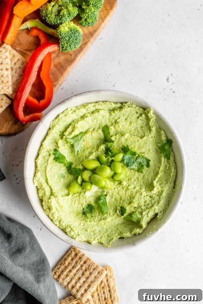A beautifully presented bowl of edamame hummus, garnished with cilantro and whole edamame beans. Slices of colorful bell pepper and crackers are arranged beside it.