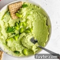 A spoon and cracker scooping edamame hummus out of a small bowl sitting on a white surface.