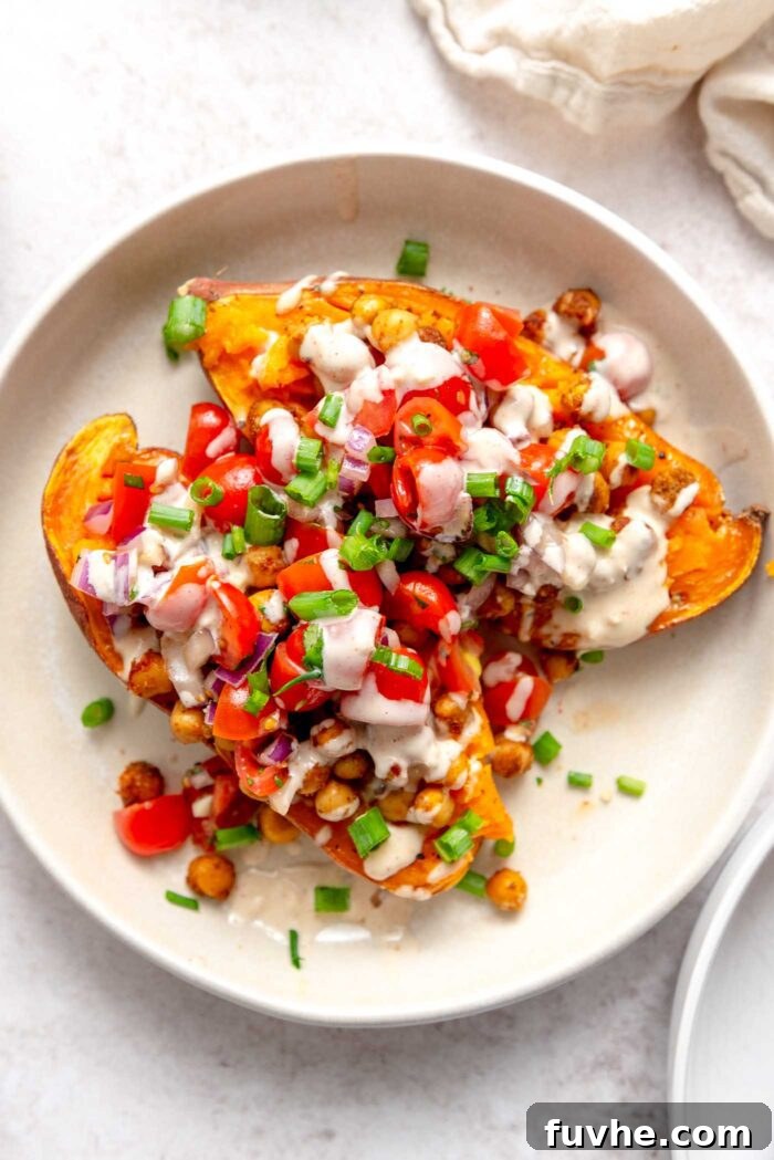 Two halves of a baked sweet potato on a plate, generously topped with roasted chickpeas, marinated tomatoes, fresh parsley, and drizzled with creamy tahini sauce.