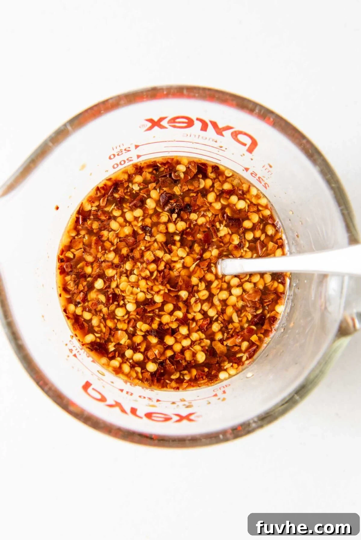 A measuring cup filled with the mixed Kung Pao sauce, featuring visible red pepper flakes, with a spoon resting inside.