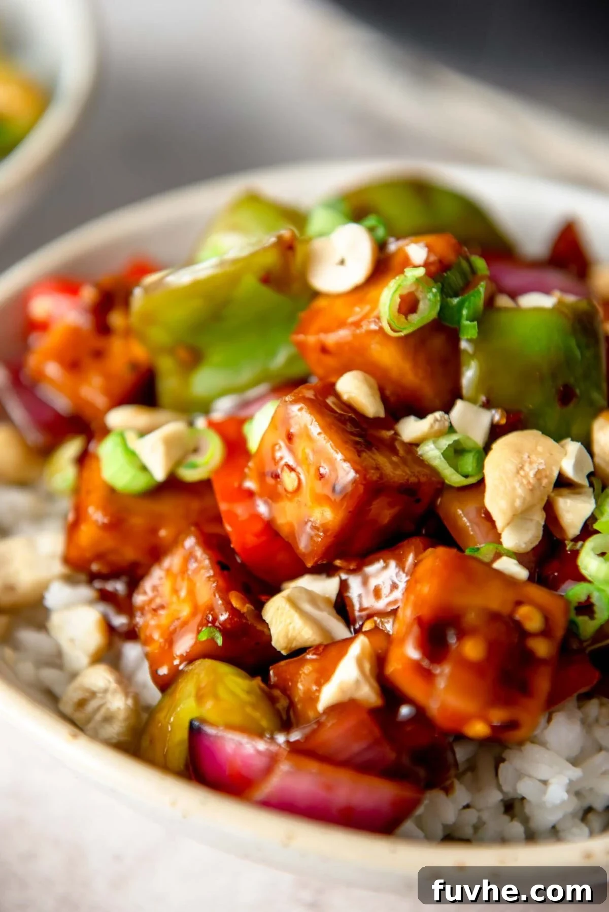 A beautifully plated bowl of Vegan Kung Pao Tofu, featuring crispy tofu and colorful vegetables in a thick sauce, served over white rice and garnished with green onions and peanuts.