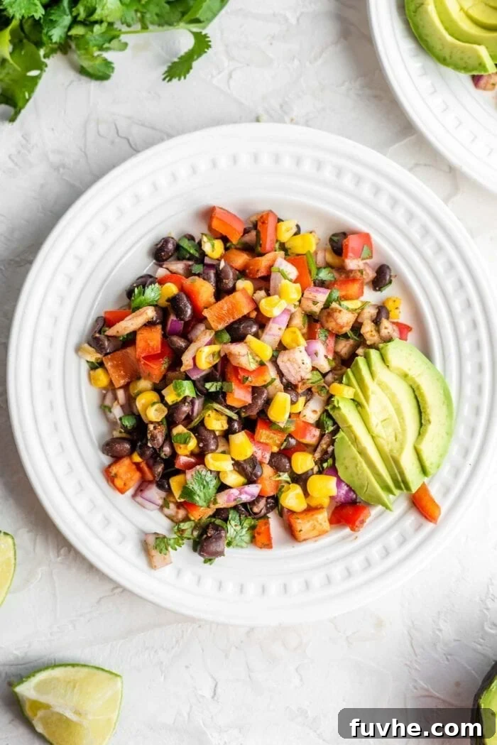 Fiesta Black Bean and Corn Salad 2 A small white plate showcasing a vibrant Southwest Black Bean Corn Salad generously topped with creamy avocado slices, garnished with fresh cilantro and a wedge of lime on the side, ready to be enjoyed.
