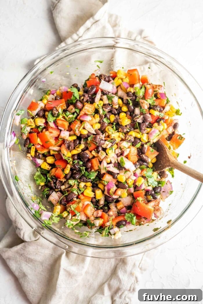 Fiesta Black Bean and Corn Salad 3 A large glass mixing bowl filled with freshly prepared Southwest black bean corn salad, featuring visible kernels of corn, black beans, diced bell peppers, and cilantro, set on a clean white surface.