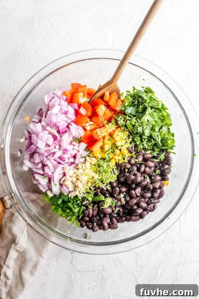 Fiesta Black Bean and Corn Salad 7 A glass mixing bowl filled with the colorful ingredients for Southwest black bean corn salad, including black beans, red onion, lime zest, minced garlic, diced bell pepper, corn, and jalapeño, before mixing.