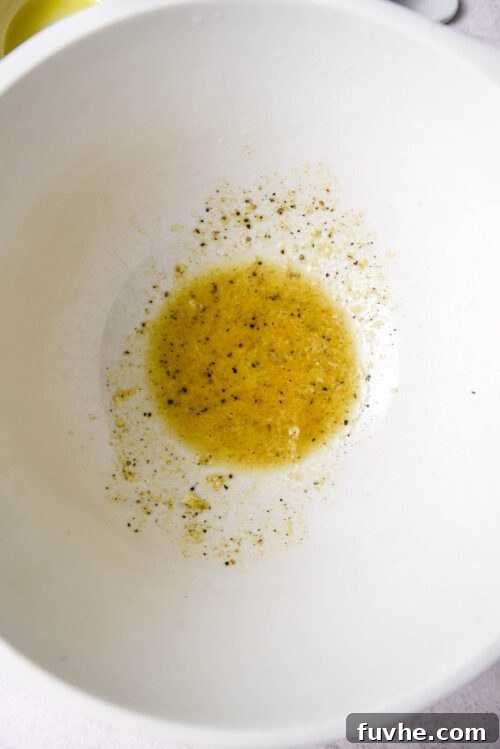 A glass mixing bowl filled with the ingredients for the olive oil vinaigrette: olive oil, lime juice, lime zest, minced garlic, and spices, ready to be whisked.