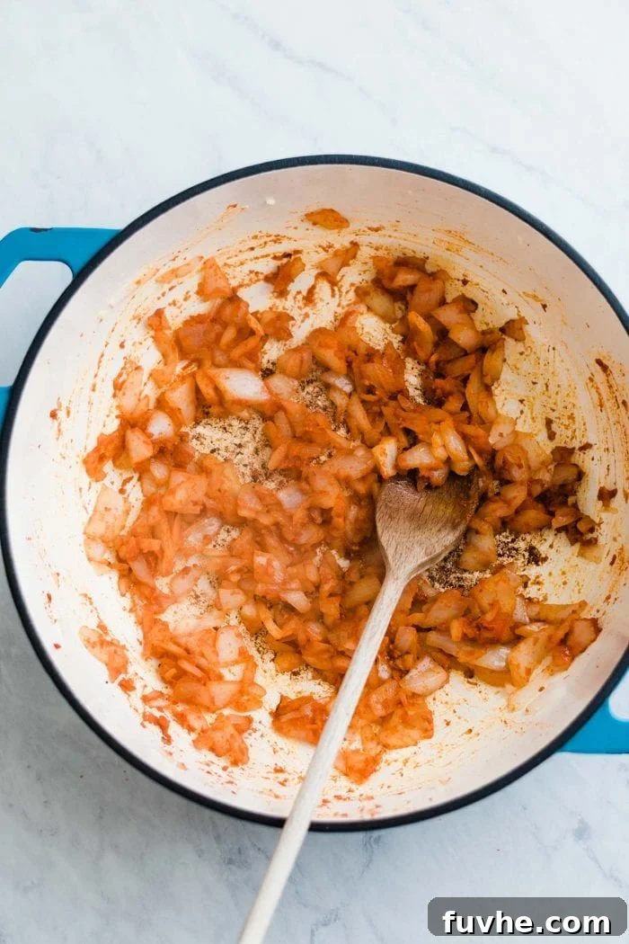 Sautéed onions, garlic, and ginger being mixed with red curry paste in a large saucepan using a wooden spoon.