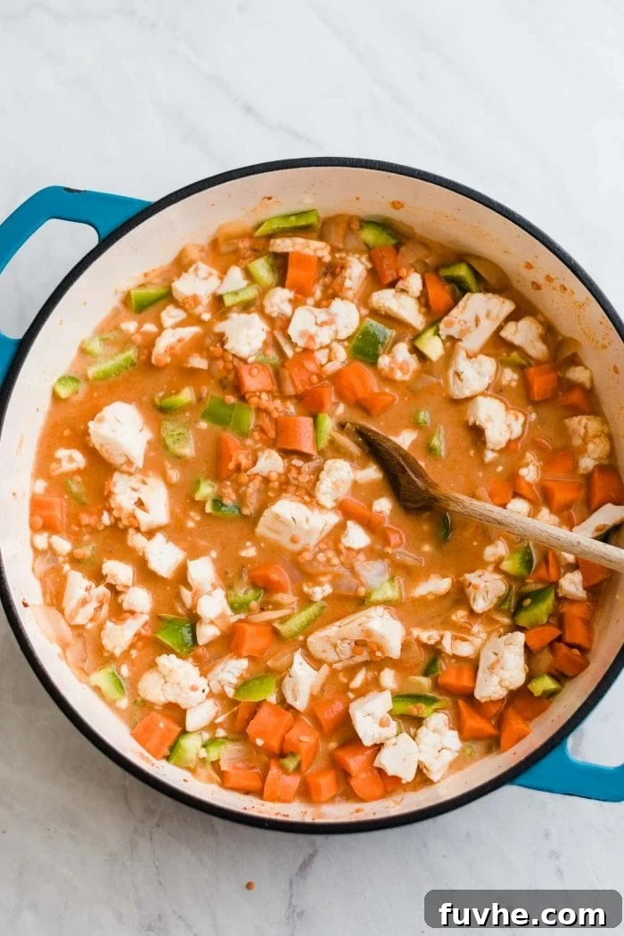 Chopped carrots, cauliflower, and green pepper simmering with red lentils in a creamy red curry broth in a large pot. A wooden spoon rests in the pot.