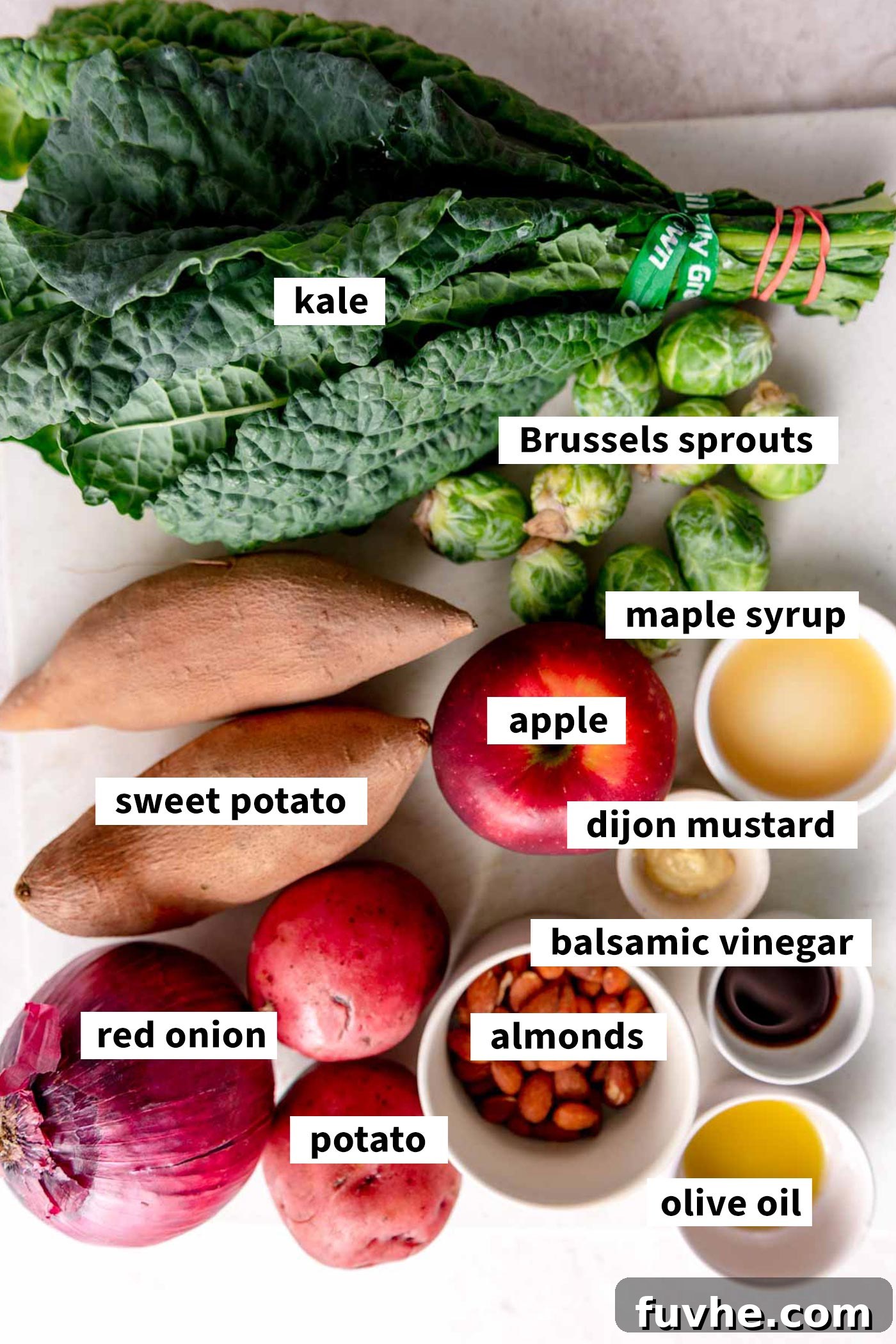 A vibrant display of fresh ingredients for an apple and kale roasted vegetable salad, including sweet potato, Brussels sprouts, red onion, kale, apple, almonds, and balsamic dressing components.