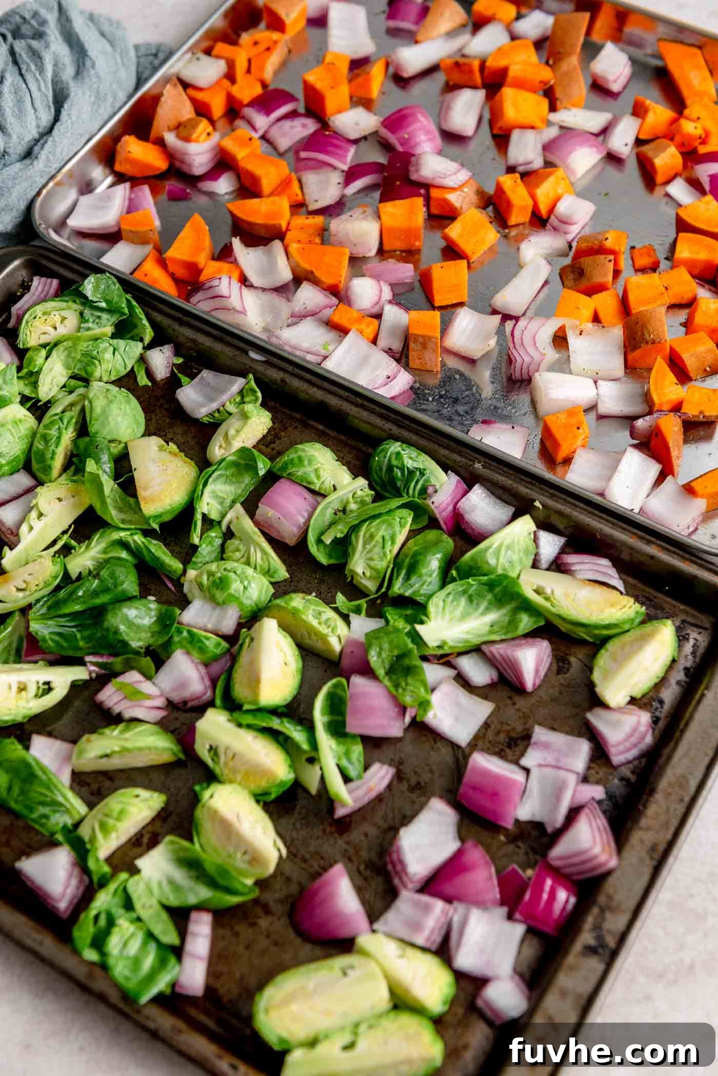 Two large baking sheets, each containing an evenly spread single layer of chopped Brussels sprouts, sweet potato, and red onion, seasoned and ready for roasting.