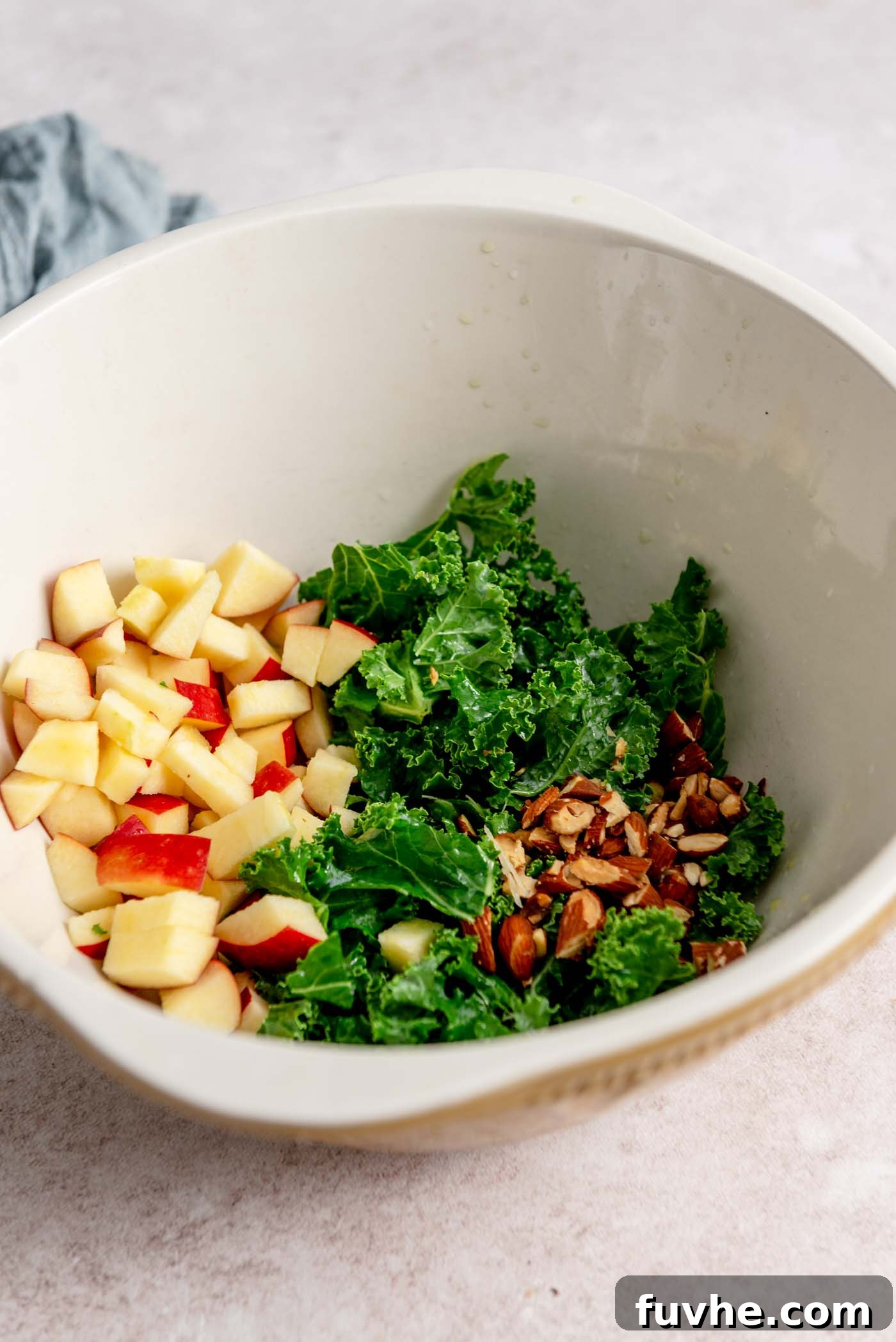 A large mixing bowl containing finely chopped kale, freshly diced apple, and crunchy chopped almonds, ready for the next step of salad assembly.