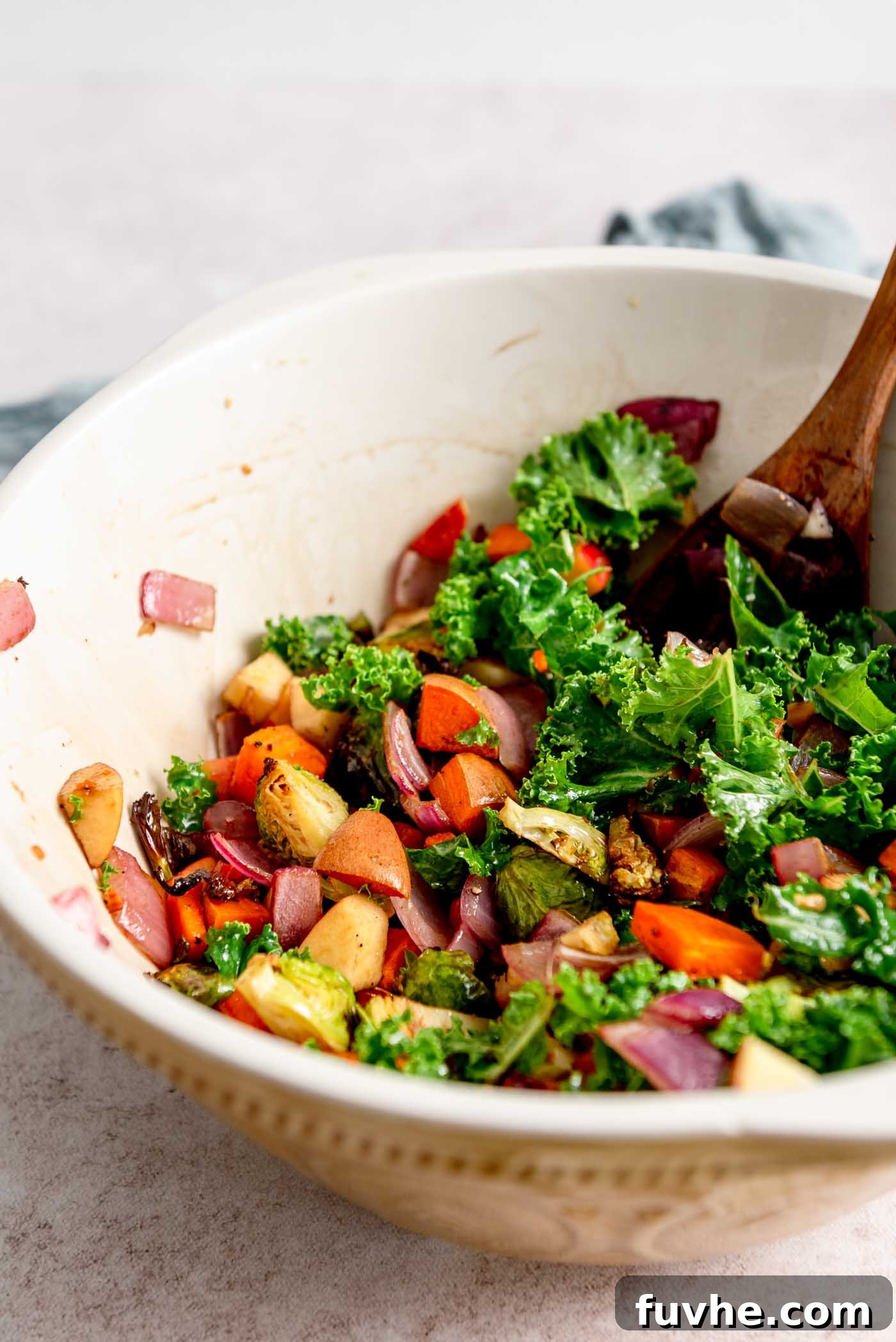A large mixing bowl brimming with the fully assembled roasted vegetable kale salad, including sweet potato, apple, and almonds, being tossed with a rustic wooden spoon.