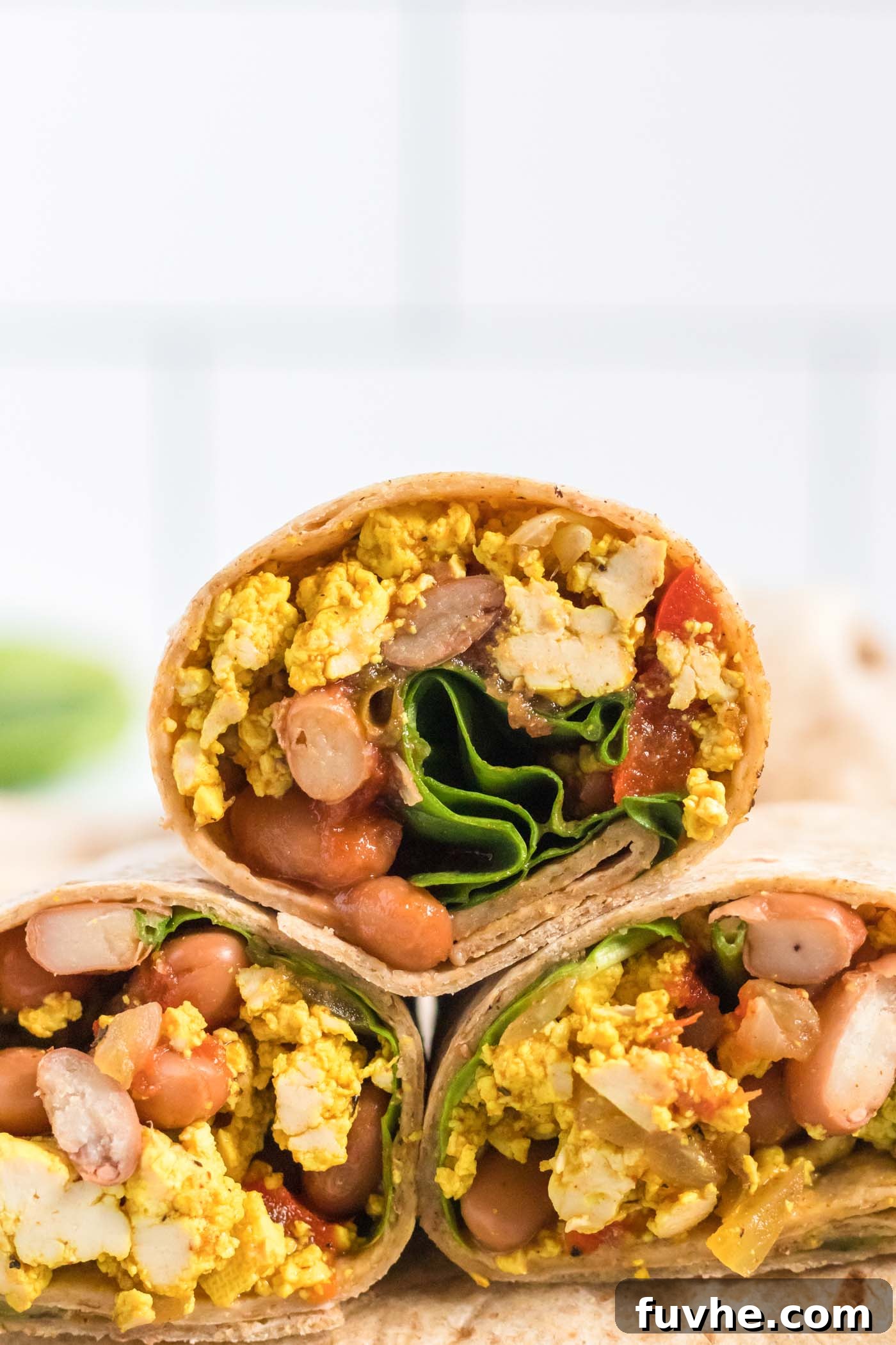 A vibrant, close-up shot of a vegan breakfast burrito sliced in half, revealing a fluffy tofu scramble, dark beans, and fresh green avocado and cilantro inside. The fillings are colorful and inviting, promising a hearty and delicious meal. The tortilla is perfectly golden brown.