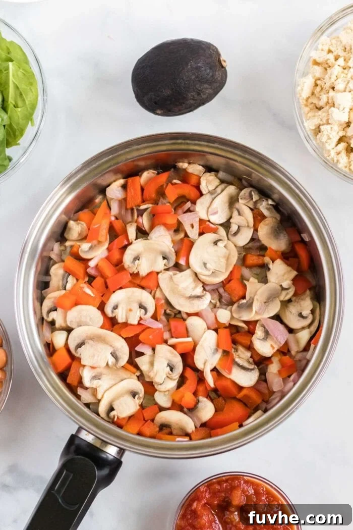 A close-up of a pan containing chopped mushrooms and red bell pepper sautéing, with an avocado and a small bowl of salsa positioned beside it. This image highlights fresh ingredients being prepared for the breakfast burrito filling.