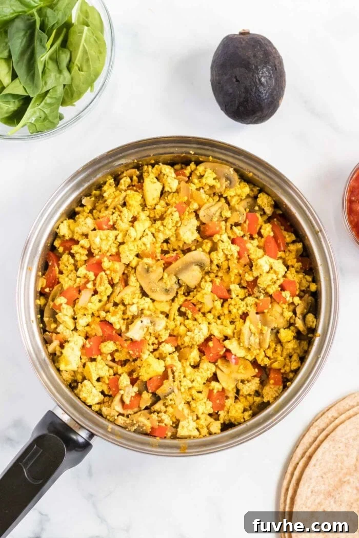A close-up shot of a savory tofu and vegetable scramble cooking in a hot pan, showing the crumbled tofu mixed with colorful bell peppers, mushrooms, and spices. Steam rises from the pan, indicating it's freshly cooked and ready to be incorporated into burritos.