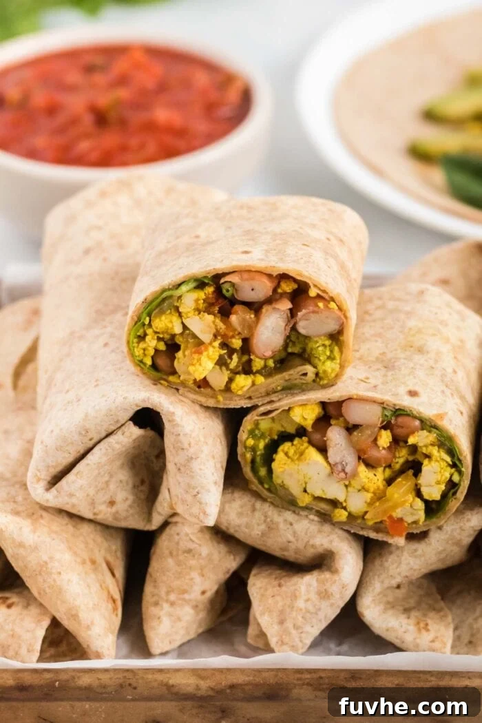 A mouth-watering vegan breakfast burrito, perfectly sliced in half to reveal its rich, vibrant fillings, rests atop a stack of additional burritos, highlighting their appeal as a delicious and satisfying meal. The presentation suggests convenience and deliciousness for meal prep.