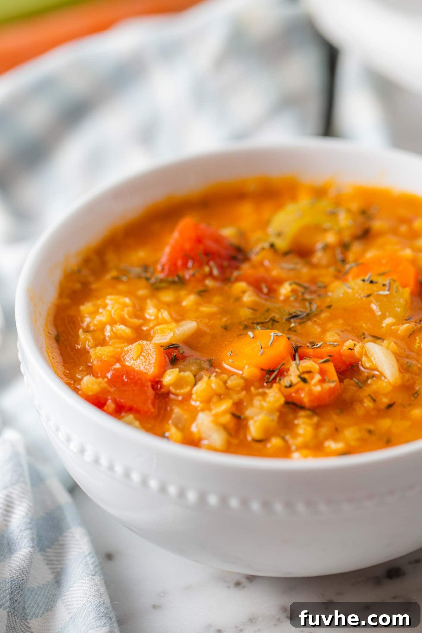 A steaming bowl of vibrant vegan red lentil tomato soup, garnished with fresh herbs and a sprinkle of black pepper. Visible bits of diced tomato, carrots, and celery add texture.