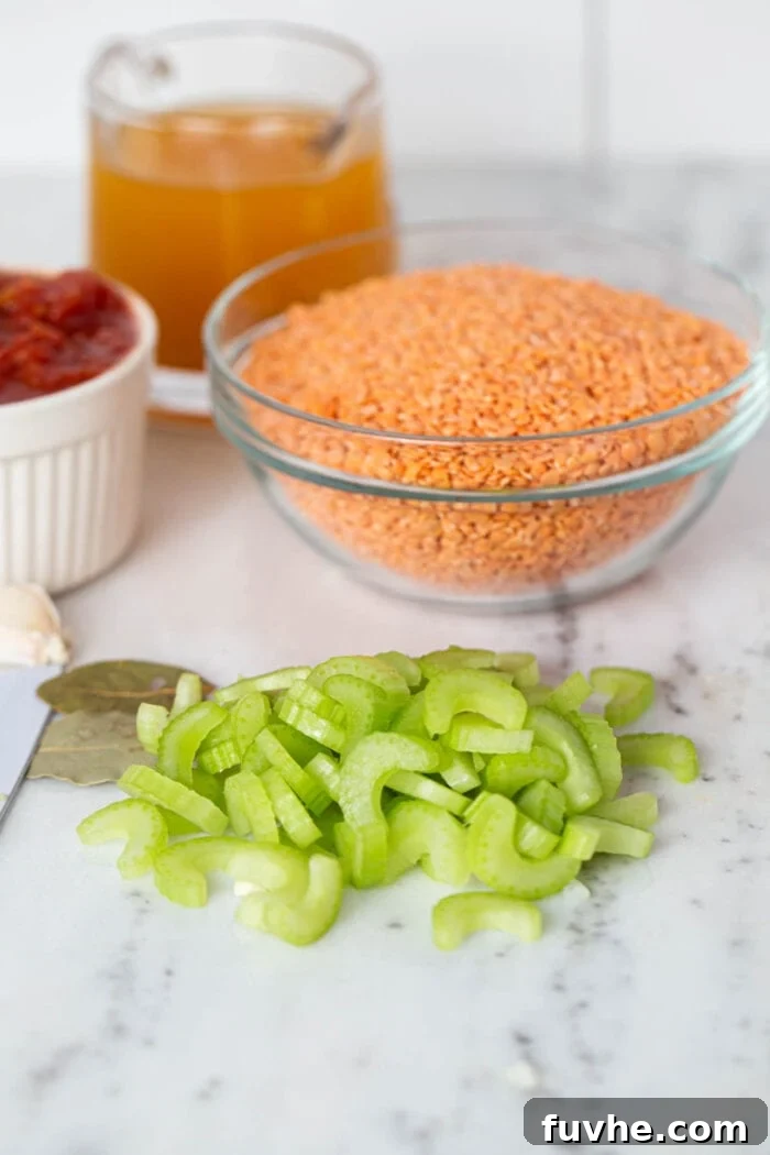 A close-up shot of freshly chopped celery on a marble countertop, ready for cooking. In the background, bowls hold pre-measured red lentils, vegetable broth, and diced tomatoes, highlighting the organized preparation for the soup.