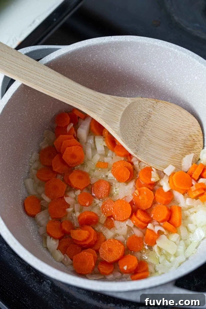 A vibrant mix of diced onions, carrots, and minced garlic gently simmering in a large soup pot, stirred by a wooden spoon. Steam rises, indicating the aromatic vegetables are cooking down to form the flavorful base of the soup.