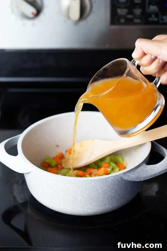 A hand carefully pouring vegetable stock into a large pot already filled with a simmering mixture of diced tomatoes and red lentils on a stovetop, capturing the moment the liquid base is added to the soup.