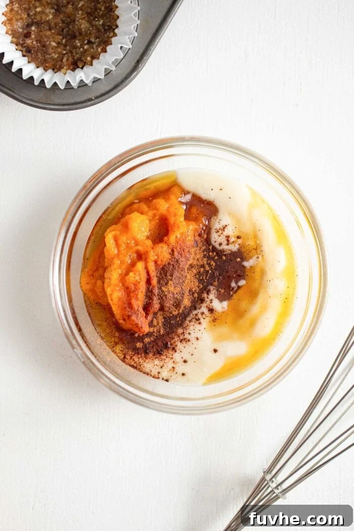 An overhead view of a small glass mixing bowl filled with smooth pumpkin puree, melted coconut butter, and a blend of aromatic spices. A whisk is positioned neatly beside the bowl.