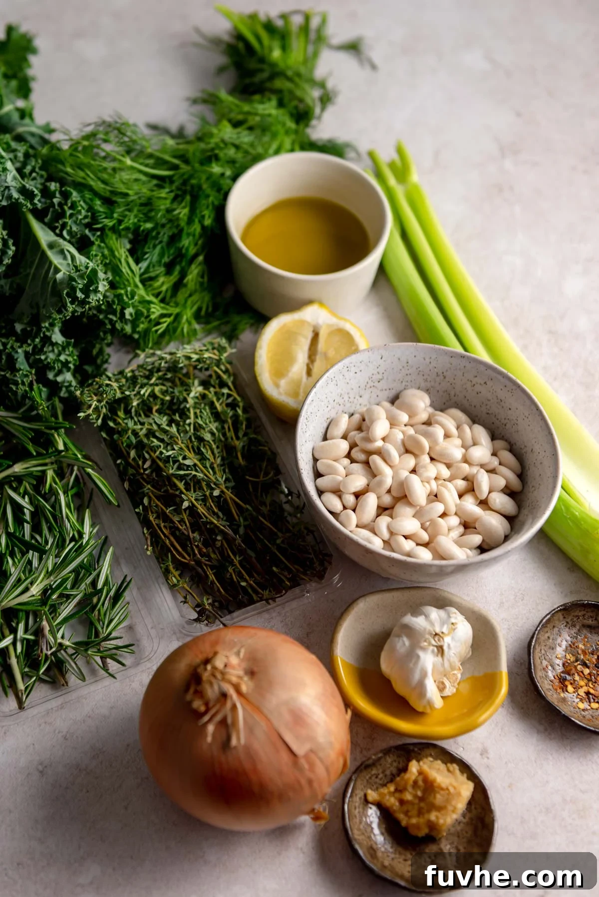 Savory Simmered Beans 3 A curated selection of fresh ingredients laid out on a countertop, including a golden onion, vibrant celery stalks, a bowl of dry white beans, a bottle of olive oil, a bright lemon wedge, aromatic garlic cloves, and sprigs of fresh rosemary and thyme.