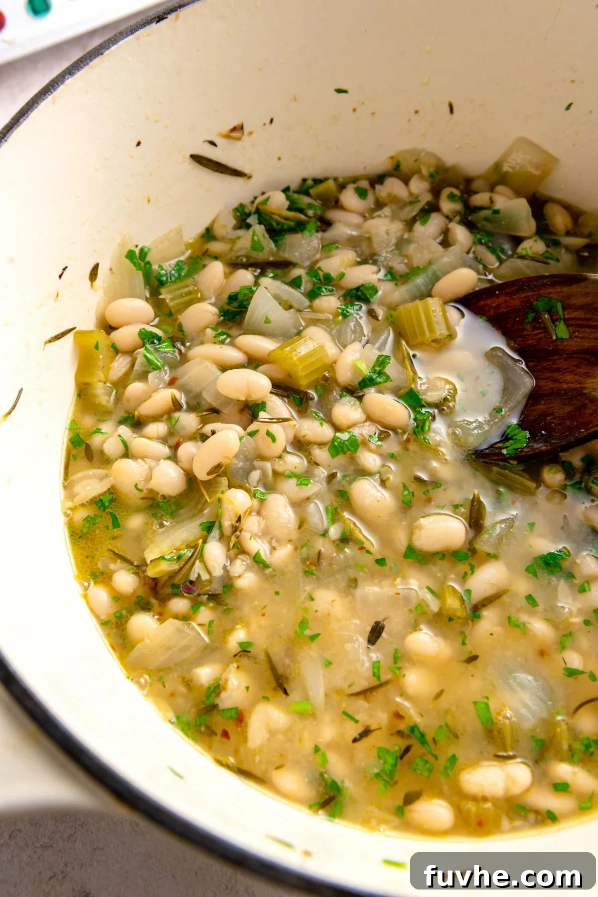 Savory Simmered Beans 4 A large pot filled with an abundance of brothy white beans, visibly infused with sprigs of fresh rosemary and thyme, and tender pieces of onion and celery, simmering to perfection.