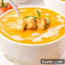 A bowl of creamy tomato soup drizzled with cream and topped with croutons and a piece of fresh basil.