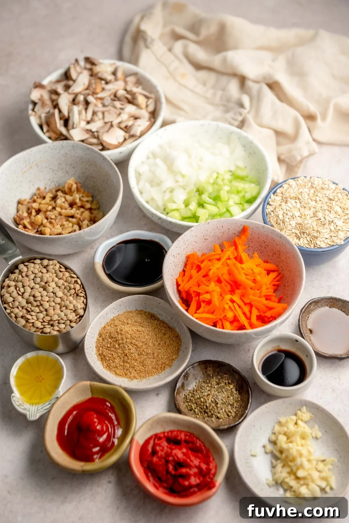 Individual bowls containing fresh ingredients for making lentil loaf, including diced carrots, walnuts, chopped celery and onion, rolled oats, sliced mushrooms, ground flaxseeds, tomato paste, ketchup, vinegar, soy sauce, and minced garlic.