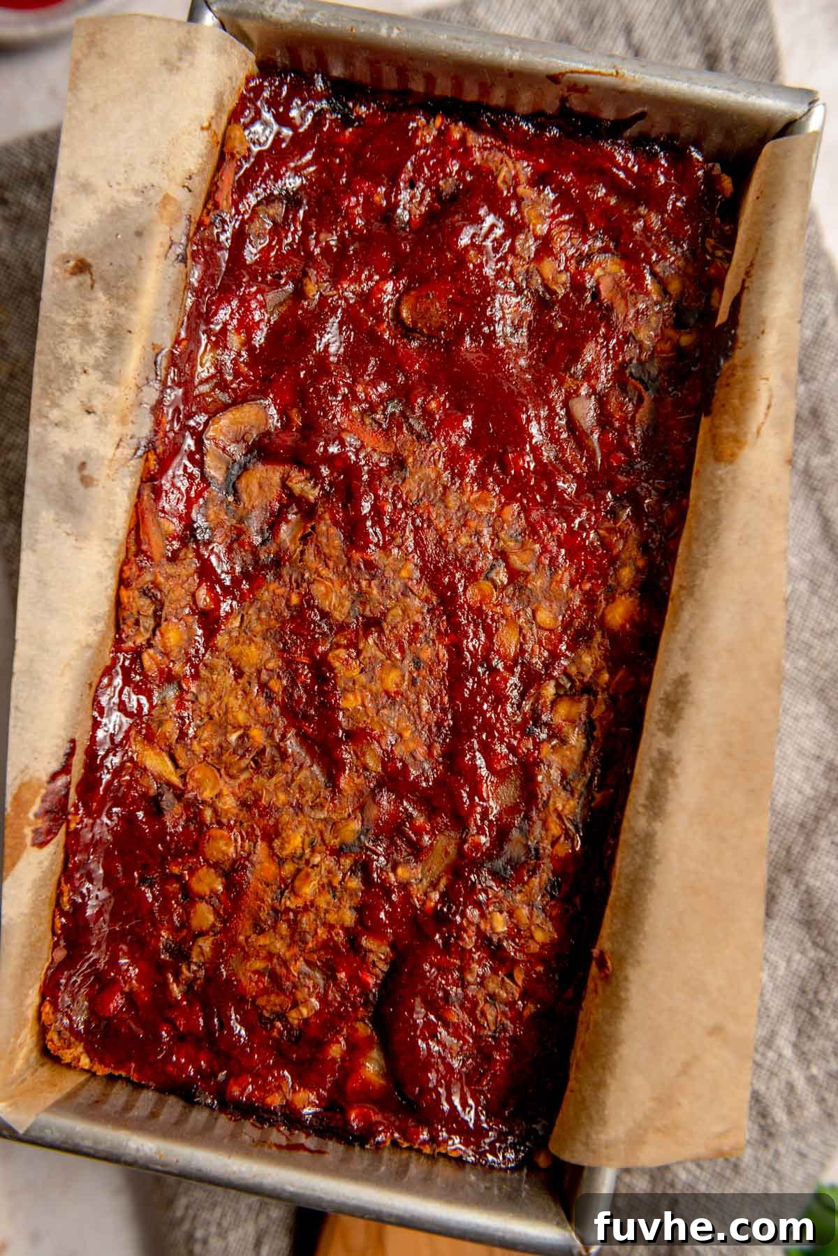 A freshly prepared lentil loaf topped with a shiny ketchup glaze, nestled in a parchment paper-lined loaf pan, ready for baking.