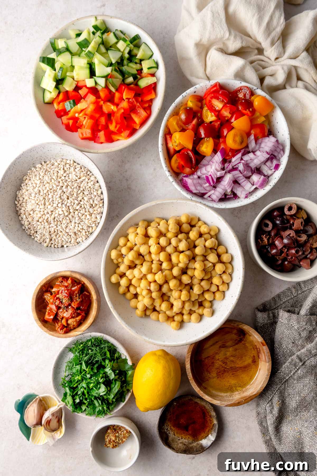 All the vibrant ingredients for making Mediterranean barley salad meticulously arranged in separate bowls on a wooden surface, including cooked barley, drained chickpeas, chopped cucumber, bell pepper, cherry tomatoes, red onion, Kalamata olives, sun-dried tomatoes, fresh parsley and dill, and a separate bowl for olive oil, lemon juice, red wine vinegar, garlic, and Dijon mustard.