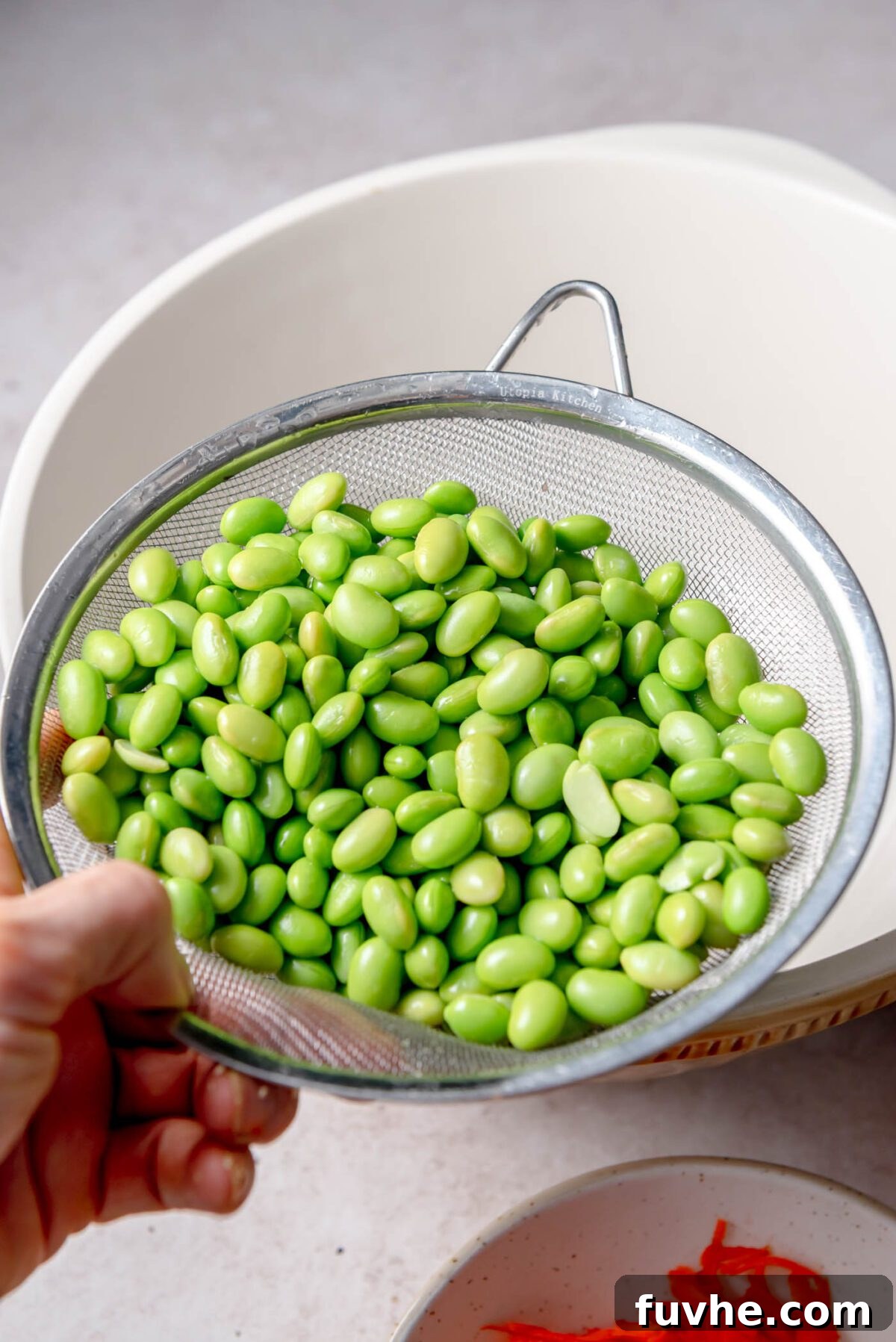 Vibrant Edamame Medley 4 A metal strainer brimming with freshly rinsed, bright green edamame beans, suspended over a large white mixing bowl, poised for incorporation into the vibrant salad.