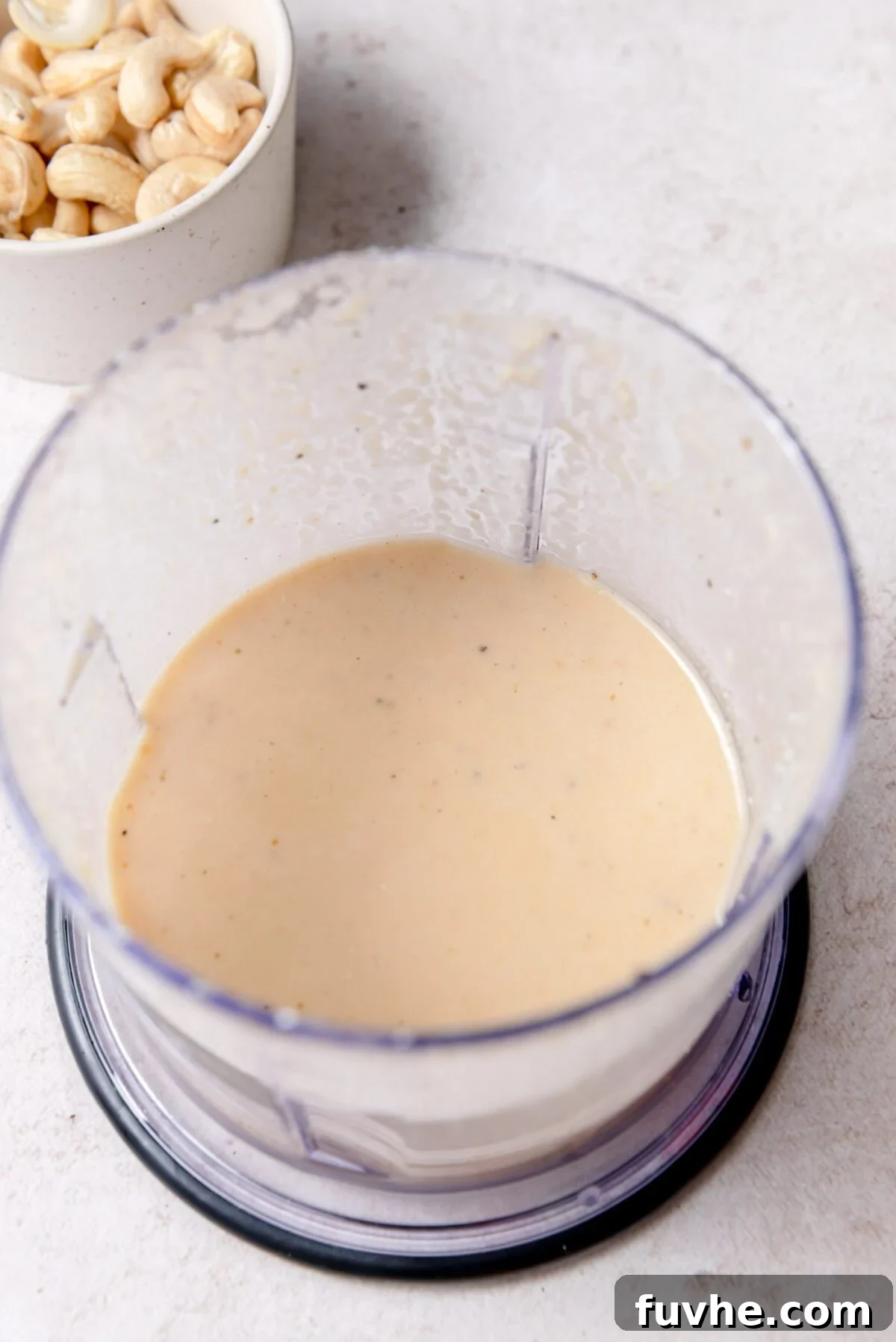 Vibrant Edamame Medley 5 A sleek blender cup containing a perfectly smooth and creamy miso ginger sesame dressing, its pale beige color flecked with tiny seasonings, resting beside a small bowl of raw cashews on a pristine countertop.