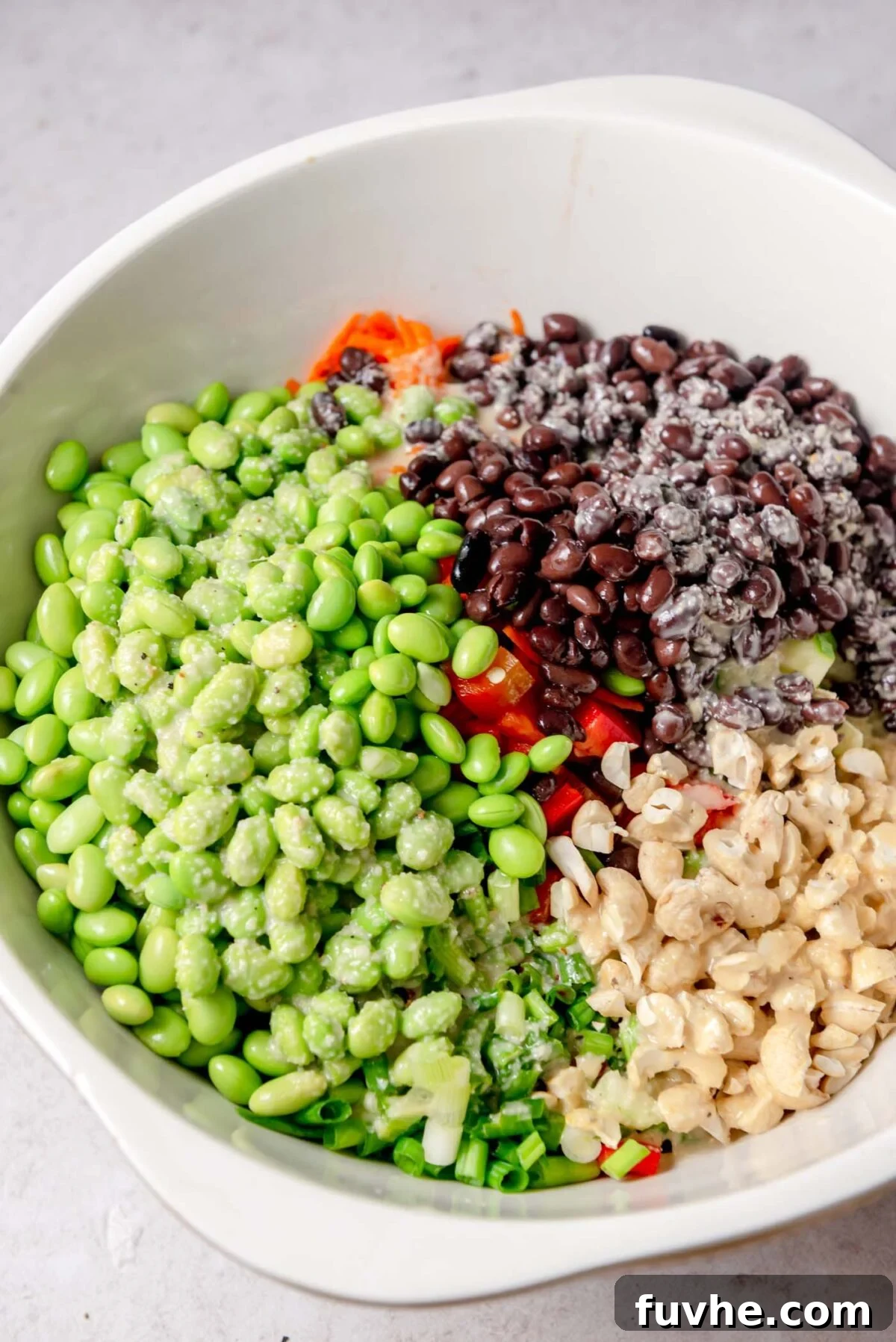 Vibrant Edamame Medley 6 A spacious white mixing bowl holds the colorful constituents of the edamame bean salad: bright edamame, dark black beans, finely diced red pepper, crunchy cashews, and thinly sliced green onions, arranged in distinct sections before being gently combined.