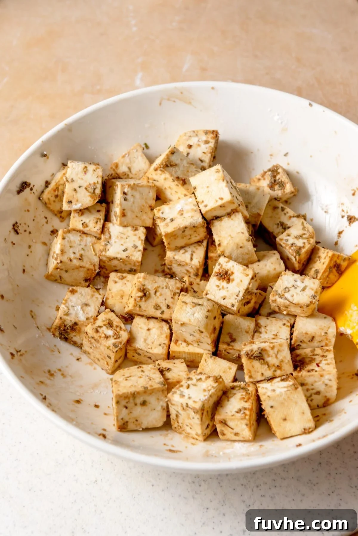 Marinated extra-firm tofu cubes tossed with a blend of herbs, soy sauce, and lemon zest in a white mixing bowl, ready for roasting to achieve crispy perfection for Mediterranean roasted veggie bowls.
