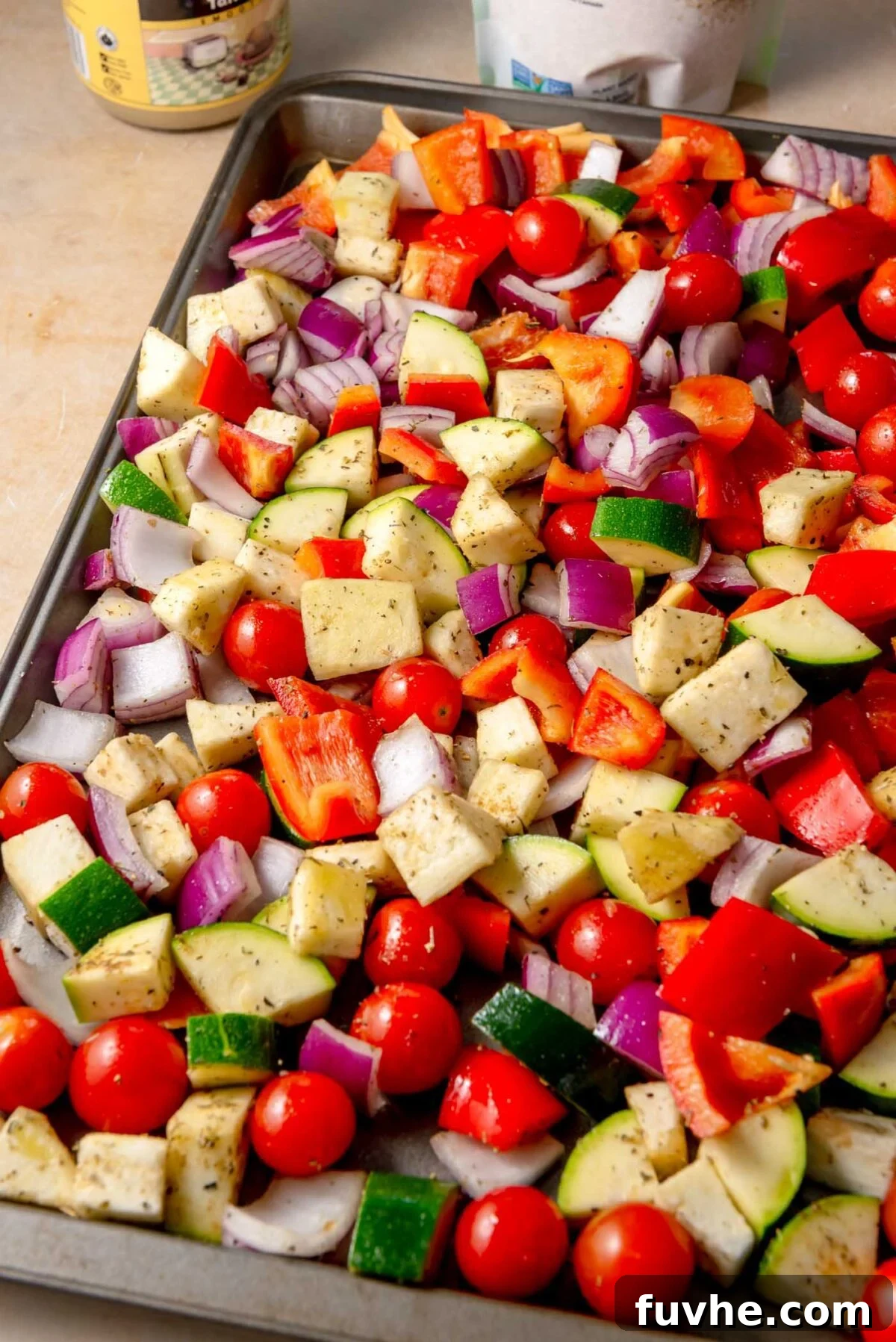A colorful medley of chopped zucchini, vibrant red bell peppers, diced eggplant, wedges of red onion, and bright cherry tomatoes spread out on a sheet pan, generously seasoned with Mediterranean herbs and olive oil, ready for roasting.