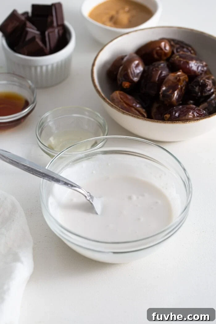 Decadent Chocolate Peanut Butter Dates 4 A small glass bowl filled with thick, white coconut cream, with a bowl of whole Medjool dates softly blurred in the background, ready for preparation.