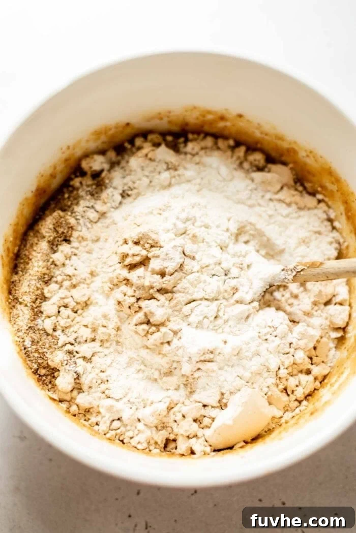 Plant-Powered Protein Muffins 5 Dry baking ingredients, including flour, oats, and protein powder, being mixed into wet ingredients in a large mixing bowl.