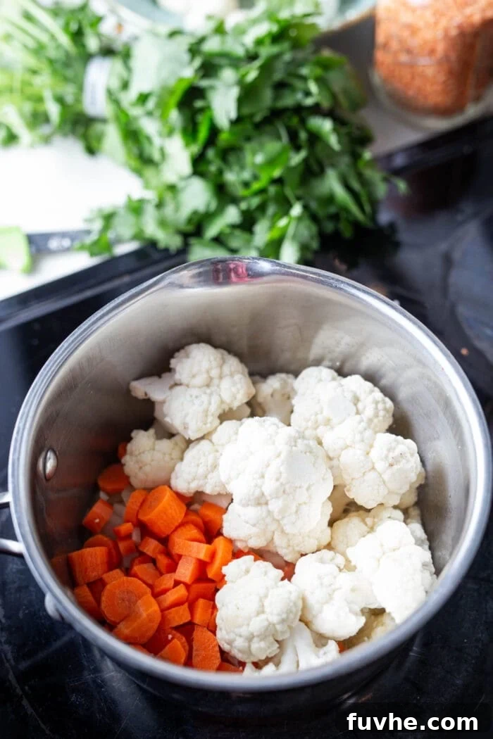 Chopped carrots, cauliflower, and red curry paste being stirred into the sautéed aromatics in a large saucepan.