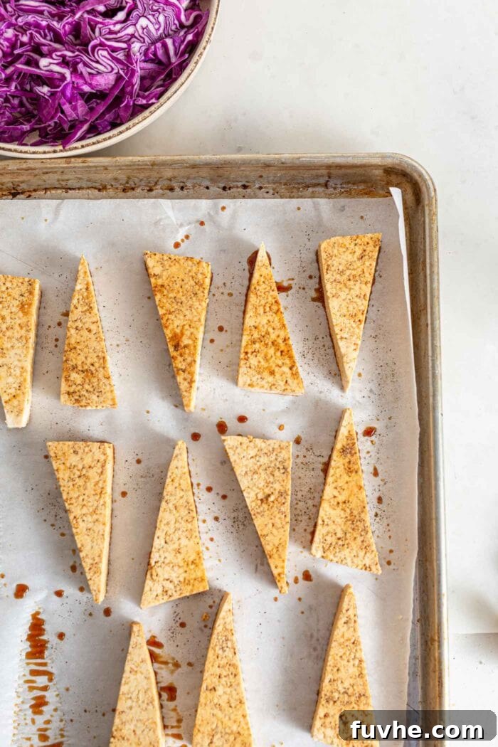 Neatly arranged sliced triangles of seasoned tofu baking on a parchment-lined baking pan, showcasing their preparation for a crispy finish.
