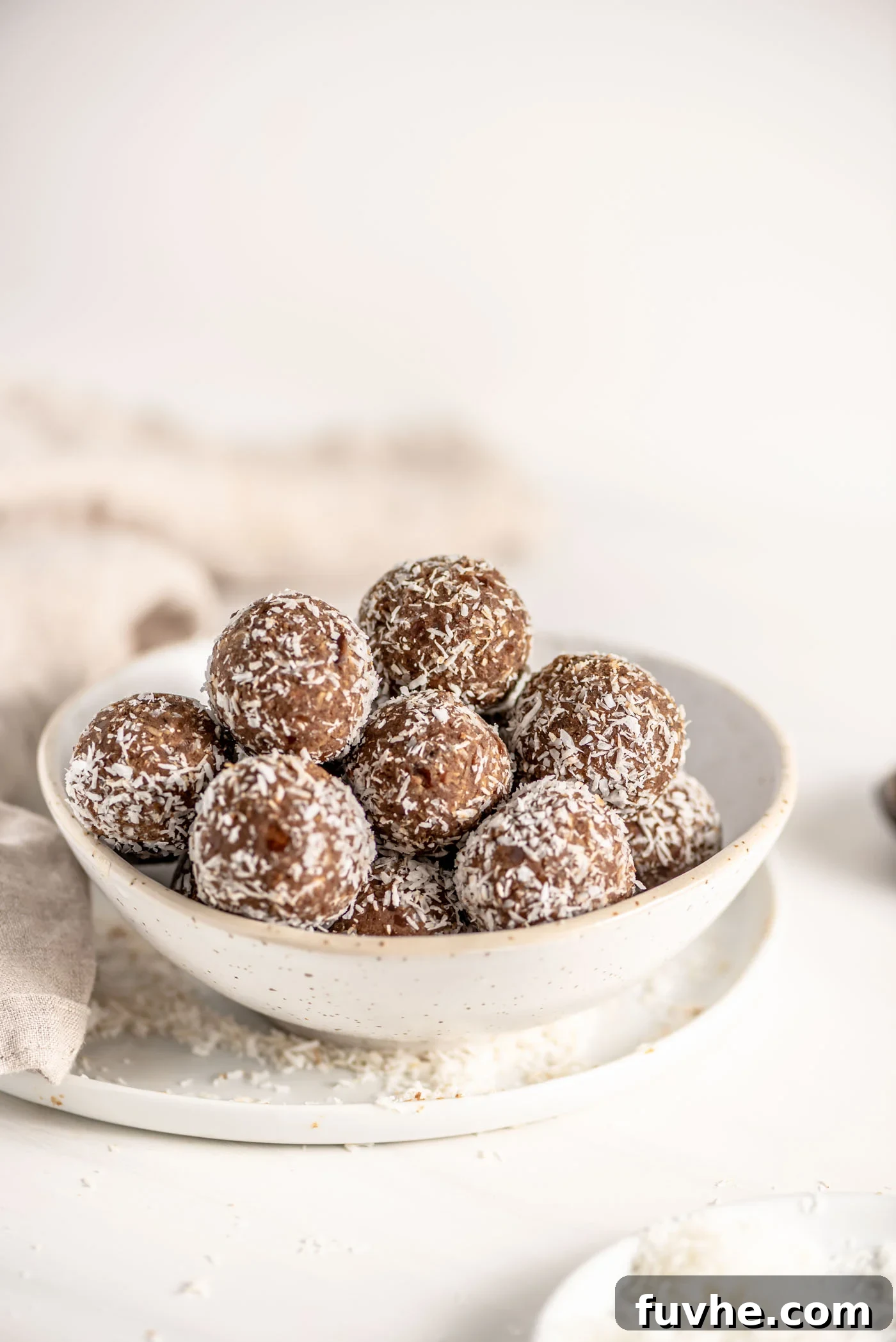 Vegan Coffee Coconut Energy Balls 2 A bowl brimming with no-bake vegan coconut coffee protein balls, generously rolled in shredded coconut, rests on a rustic plate. A soft dishcloth provides a cozy background.