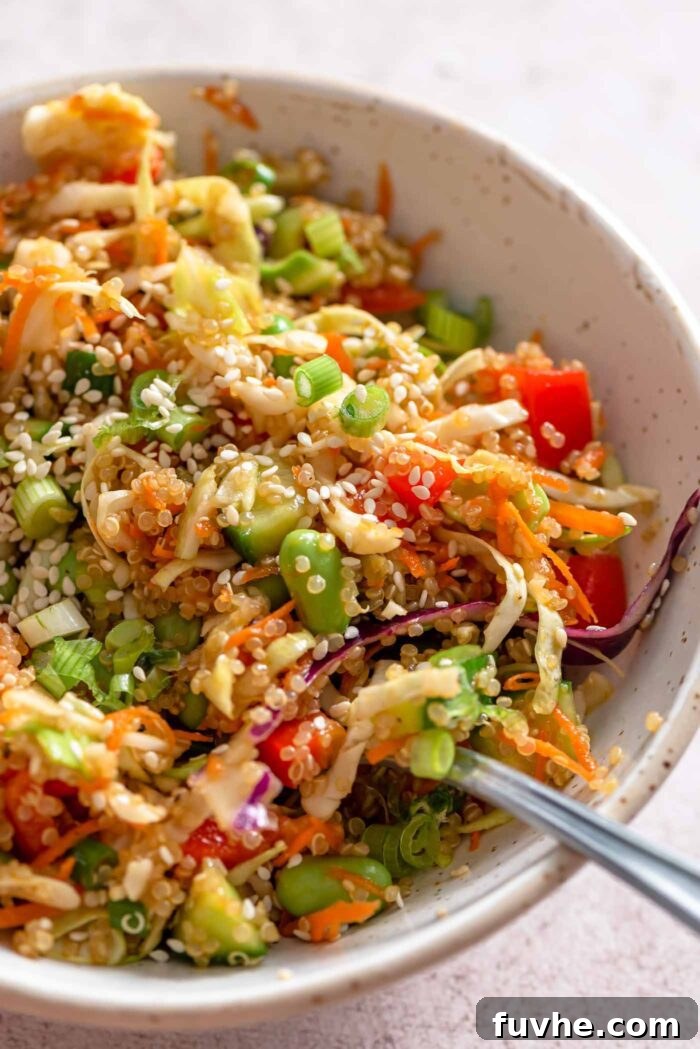 A fork resting in a small bowl of quinoa edamame salad with cabbage, green onion, red pepper, carrot and sesame seeds. 