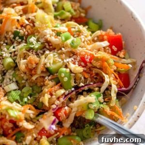 A fork resting in a small bowl of quinoa edamame salad with cabbage, green onion, red pepper, carrot and sesame seeds.