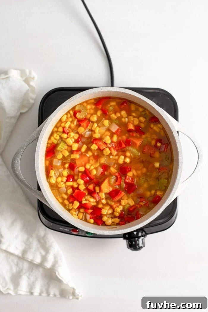 Unblended sweet potato corn chowder, rich with colorful pieces of bell pepper, corn, sweet potato, celery, and onion, gently cooking in a soup pot on a small cooktop.