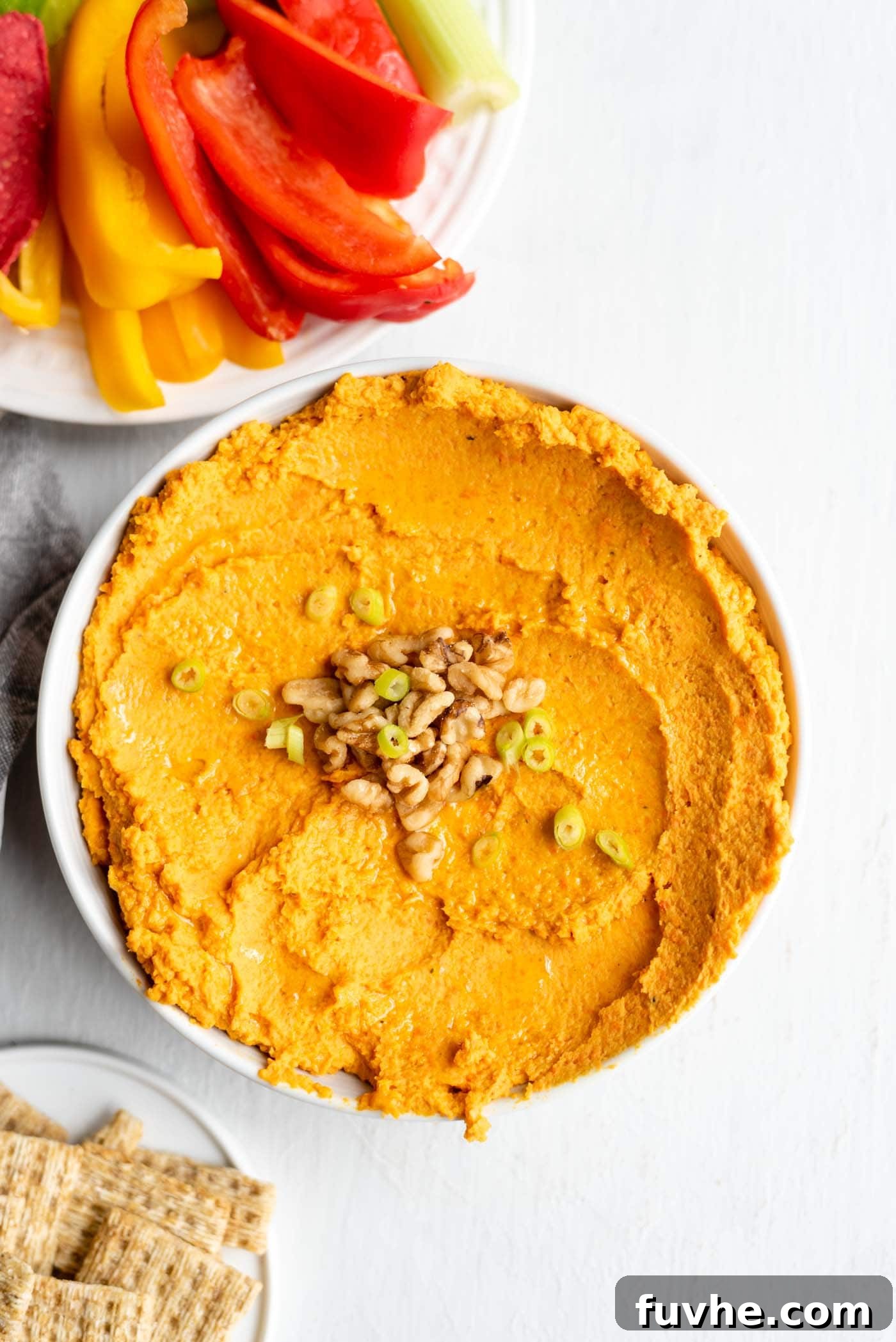 A bowl of roasted carrot hummus topped with walnuts and fresh green onion, served alongside an assortment of crisp crackers and fresh vegetable sticks, highlighting its rich color and inviting texture.