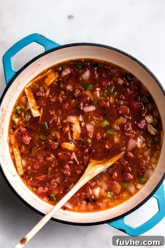 Vegan tortilla soup simmering gently in a blue soup pot, stirred with a wooden spoon, showcasing its rich, developing texture.