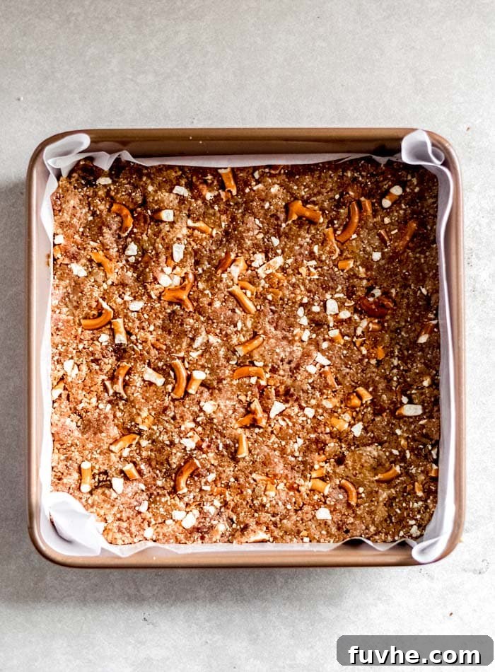 Neatly pressed pretzel granola bar mixture in a square pan, lined with parchment paper, preparing for the chilling process.
