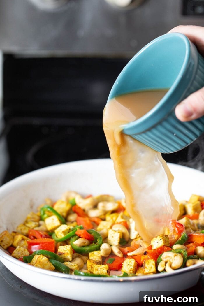 Crispy Cashew Tofu Fried Rice 5 A stream of vegetable broth mixture being poured into a hot skillet, bubbling around the cooked tofu, cashews, and sautéed vegetables.