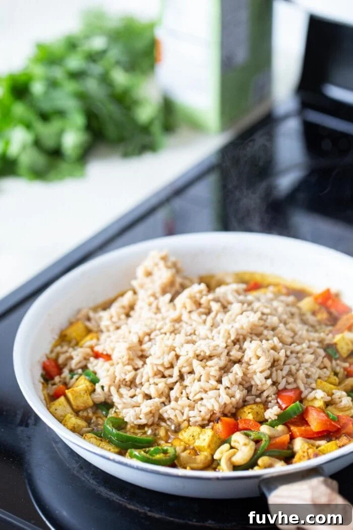 Crispy Cashew Tofu Fried Rice 6 Fluffy, cooked brown rice being stirred into a vibrant medley of tofu, vegetables, and cashews in a large skillet, transforming into flavorful fried rice.